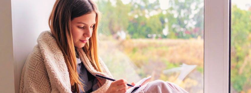 Woman sitting by window while wrapped in a blanket and journaling, representing self-compassion and the understanding that early coping adaptations were brilliant solutions to a difficult environment